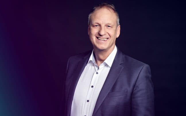 Professionelles Foto von einem Trainer. Er trägt einen Anzug und eine Brille. Er lächelt in die Kamera.